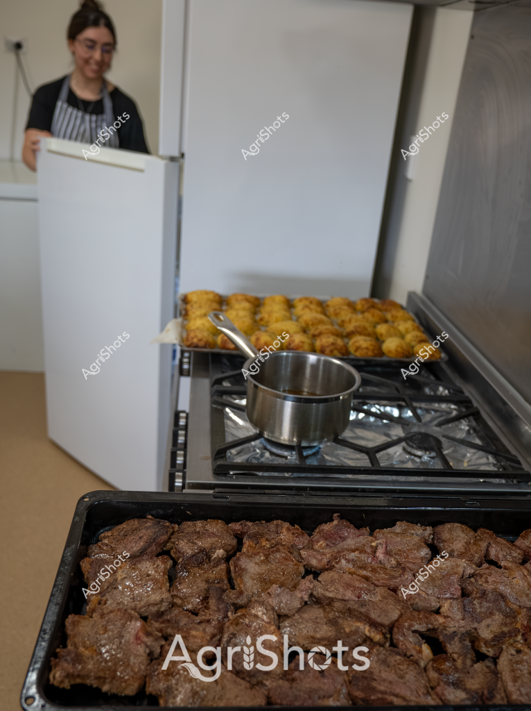 A woman in a black and white striped apron stands in a commercial kitchen, preparing a batch of roasted meats and golden-brown potato dishes. In the foreground, a tray of richly browned meat cuts sits ready for serving, while another tray of crispy, golden potato items is positioned on the stovetop. A stainless steel pot rests on the gas stove, suggesting ongoing food preparation. The kitchen environment appears clean and professional, with white walls and stainless steel equipment, indicating a carefully managed food production setting. The scene captures the precision and care involved in commercial food preparation, with each element meticulously arranged and cooked to perfection.