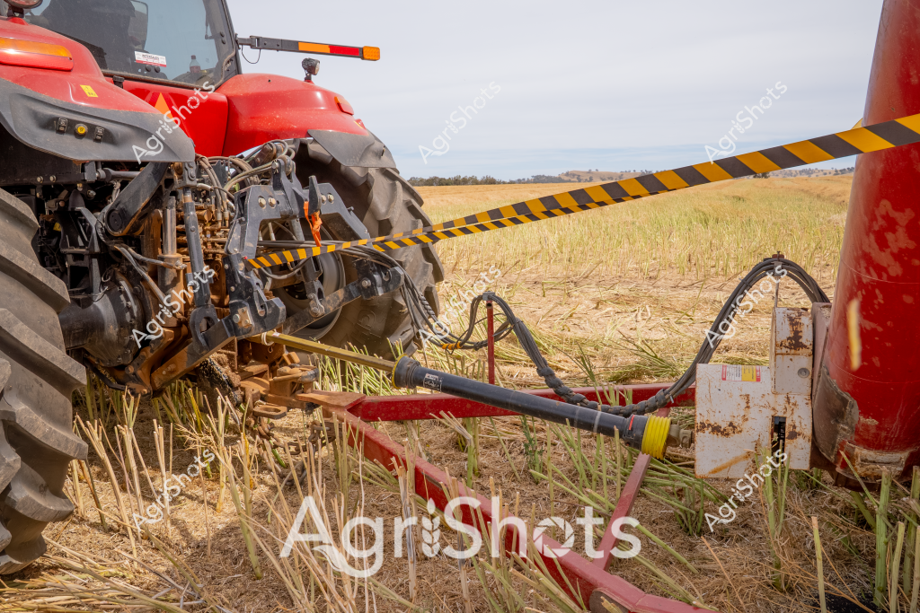 A red tractor positioned in a stubble field, showcasing sophisticated agricultural machinery connections. Intricate hydraulic linkages and robust mechanical attachments are visible, connecting the powerful tractor to an agricultural implement. Yellow and black caution tape-styled markings highlight critical connection points, emphasizing safety and precision in farm equipment integration. The landscape behind reveals a golden-hued harvested field with remnant crop stubble, stretching toward a distant horizon under an overcast sky. Large agricultural tires with deep treads grip the uneven ground, demonstrating the heavy-duty design required for modern farming operations. This image captures the technical complexity of contemporary agricultural machinery, where engineering precision meets rural productivity.
