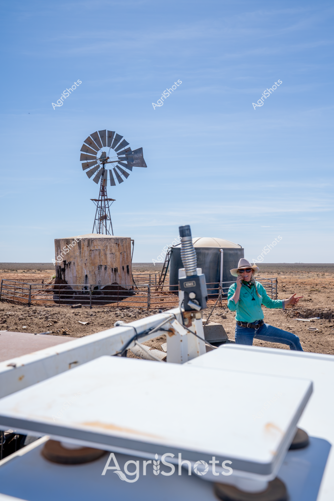 A woman in a turquoise shirt and wide-brimmed hat stands amid a stark, arid landscape, conducting agricultural field research or survey work near a classic windmill and water storage tank. The windmill, a quintessential feature of Australian rural infrastructure, rises against a clear blue sky, its metal blades poised to catch the slightest breeze. A white instrument or measurement device is mounted in the foreground, suggesting scientific data collection or environmental monitoring. The surrounding terrain is a palette of browns and tans - dry, compacted earth characteristic of drought-affected or semi-arid regions of Australia. This scene captures the resilience of agricultural workers and the essential infrastructure that supports farming in challenging environmental conditions, where water management and precise environmental measurements are critical to survival and productivity.