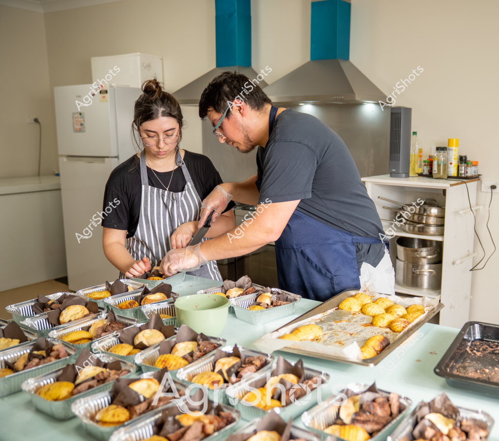 Two people prepare a large batch of packaged meals in a commercial kitchen setting. A woman in a striped apron and a man in a gray t-shirt are carefully portioning and packaging food items into aluminum trays. The kitchen workspace is covered with numerous prepared meal containers, featuring yellow-golden baked items and what appear to be meat portions. They are working collaboratively, with precise and methodical movements, suggesting this is a professional food preparation operation. The stainless steel kitchen environment, with a white refrigerator and cooking range in the background, indicates a commercial or catering kitchen setup. The meal trays are neatly arranged, showing a systematic approach to bulk food preparation, likely for a catering service, meal delivery program, or institutional food service.