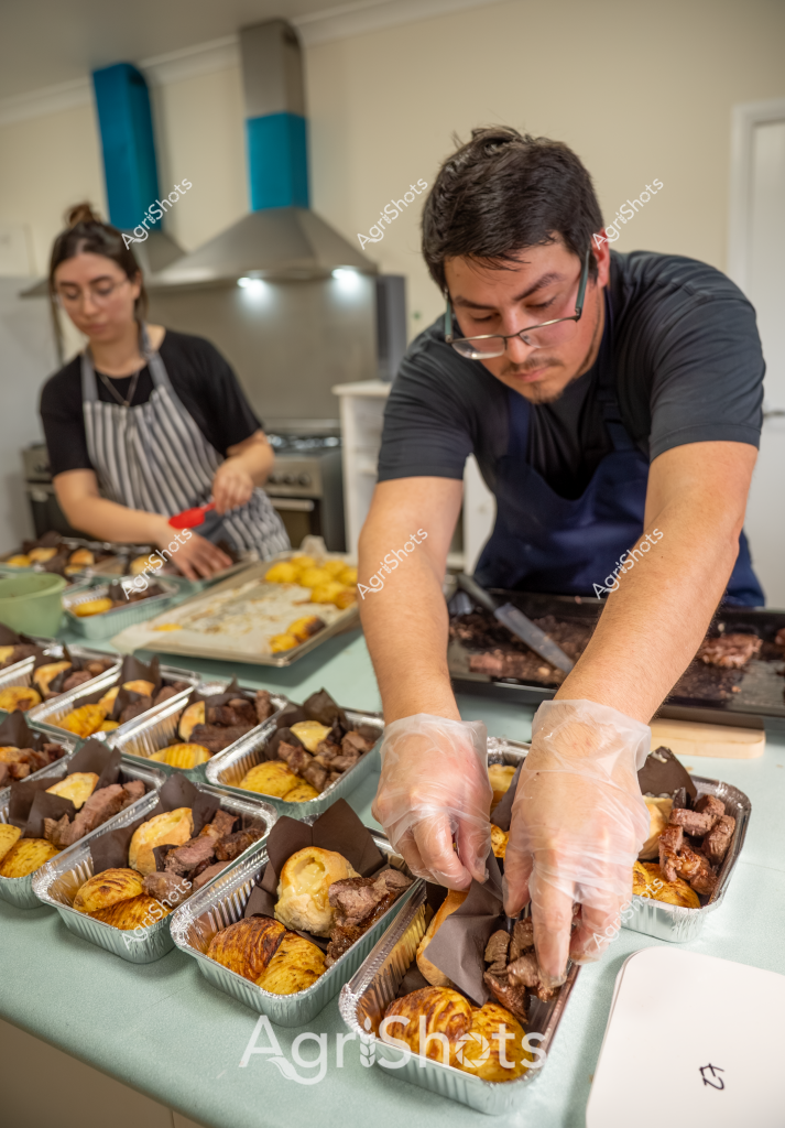 A man and a woman in dark-colored aprons and protective gloves are carefully preparing meal portions in aluminum trays in a commercial kitchen. They are meticulously arranging what appears to be meat cuts and side dishes - with golden-colored items (possibly roasted potatoes or pastries) and darker meat portions distributed into individual serving containers. The kitchen has a stainless steel range hood and the workers are focused intently on their task, wearing disposable gloves to ensure food safety and hygiene standards.

The scene looks like a professional food preparation environment, with precise portioning and packaging of what seems to be pre-prepared meals or catering services. The man is wearing glasses and concentrating closely as he places meat portions into the trays, while the woman in a striped apron is working on another section of the preparation area. The aluminum trays create a uniform, organized look to the food service process, suggesting a systematic approach to meal production.

The lighting is bright and clean, typical of a professional kitchen setting, with white walls and a modern cooking environment. Each tray contains a carefully balanced combination of proteins and accompaniments, indicating a thoughtful approach to meal composition and presentation.