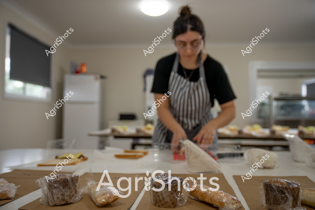 A woman baker prepares baked goods in a commercial kitchen, wearing a black t-shirt and striped apron. She stands at a white kitchen bench surrounded by freshly baked bread and pastries wrapped in clear plastic. Multiple baking trays are visible in the background, suggesting a professional bakery or commercial food preparation setting. The kitchen appears clean and well-organized, with a white refrigerator and roller blind in the background, creating a bright and efficient workspace. Her focused posture indicates careful preparation and attention to detail in her baking process.