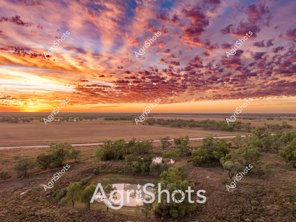 A breathtaking aerial landscape captures a dramatic sunset over an expansive rural Australian property, with an extraordinary cloudscape painting the sky in vibrant oranges, purples, and golden hues. The foreground reveals a small watering hole surrounded by scattered eucalyptus trees and sparse bushland, nestled amid broad agricultural paddocks that stretch towards a distant horizon. Strategic land management is evident in the carefully cultivated fields, which transition from dense tree clusters to open cropping areas, demonstrating the intricate balance between natural landscape and agricultural development. The sky dominates the image, with an extraordinary textured cloud formation creating a mesmerizing canvas of color that transforms the entire scene, casting warm light across the undulating terrain. Soft shadows and the gentle contours of the landscape suggest this is likely an inland region, potentially in Queensland or New South Wales, where agricultural properties often span vast distances. The watering hole provides a critical water source for the surrounding ecosystem, highlighting the importance of water management in rural Australian agricultural settings.