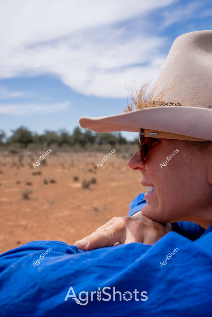 A woman stockman in a cream Akubra hat and sunglasses surveys a dry, rust-colored landscape from what appears to be an open vehicle or observation point. Wearing a bright blue shirt, she looks out across a sparse, arid paddock with scattered vegetation and distant tree lines under a soft blue sky with wispy clouds. Her profile reveals a confident, relaxed posture, suggesting extensive experience in rural Australian farming environments. The landscape hints at challenging drought conditions typical of inland Australian agricultural regions, with the woman's attentive gaze reflecting the resilience of farmers monitoring their land during difficult seasonal periods.