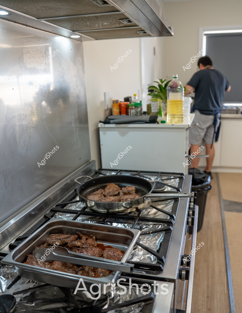 A man in a dark shirt and shorts stands at the kitchen counter, preparing a meal. On the stainless steel stovetop, multiple cuts of beef are being cooked in a frying pan and a large stainless steel tray lined with aluminum foil. The meat appears to be seared or grilled, with a rich brown color indicating thorough cooking. A large bottle of golden cooking oil sits nearby on the white kitchen counter, alongside various other cooking ingredients and a small green plant.

The kitchen is modern and clean, with a stainless steel rangehood providing ventilation over the gas stove. The cooking surface is meticulously prepared with foil, suggesting careful meal preparation. The scene captures a moment of home cooking, with precise attention to detail in the cooking process. The neutral color palette of whites, grays, and stainless steel creates a crisp, contemporary kitchen environment.

This image represents a typical Australian home cooking scene, showcasing domestic food preparation with what appears to be high-quality beef cuts being carefully prepared for a meal.