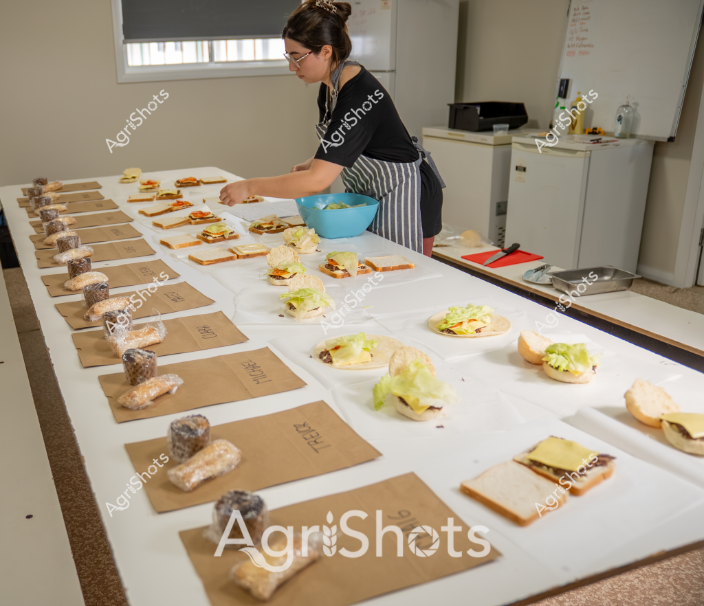 A woman in a black top and striped apron carefully prepares multiple sandwiches in what appears to be a professional food preparation area. The workspace is meticulously organized, with brown paper bags labeled and lined up on one side and an array of sandwich components systematically arranged across a white surface. A blue mixing bowl sits nearby as she arranges lettuce, bread, and various fillings with precise attention to detail.

The scene looks like a food testing or recipe development kitchen, with multiple identical sandwich variations being constructed. Each sandwich seems to be part of a controlled culinary experiment, with carefully measured ingredients and a methodical approach to assembly. The environment is clean and professional, with white walls, a window with blinds, and a neutral color palette that emphasizes the focus on food preparation.

The woman's concentration is evident as she works, positioning ingredients with care and maintaining a structured workflow. This could be a commercial kitchen, a food research facility, or a professional culinary testing environment where precision and consistency are key objectives.