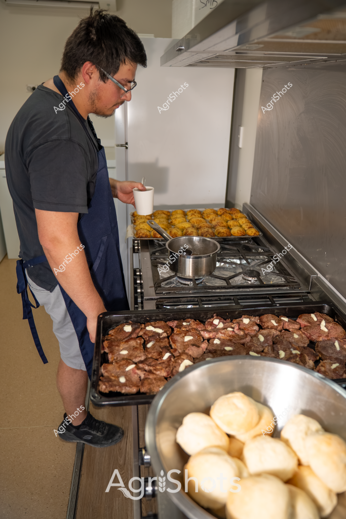 A man in a navy apron and dark polo shirt stands at a commercial kitchen stove, preparing a batch of baked goods. A large baking tray of golden-brown pastries or cookies sits on the stovetop, while another tray of rich chocolate-colored baked items is also visible. In the foreground, a metal bowl contains freshly baked round bread rolls or buns. The kitchen appears clean and professional, with stainless steel equipment and a gas stove, suggesting this is a commercial bakery or food preparation setting. The man is carefully monitoring the baking process, holding a small white container, and demonstrating careful attention to his culinary craft.