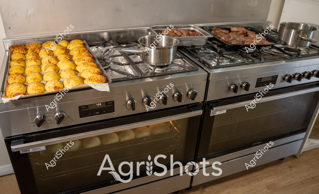 A professional stainless steel kitchen stove is laden with multiple culinary preparations - golden roasted corn on the left side, perfectly grilled meat cuts on the right, and a gleaming pot simmering on the central gas burners. The corn appears freshly baked, displaying a rich yellow-golden color with a slightly crisp exterior, neatly arranged in rows across a baking tray. Beside the corn, two trays of meat sit sizzling on the gas stovetop, their rich brown sear indicating high-heat cooking. A stainless steel pot sits centrally, suggesting ongoing meal preparation. The professional-grade oven's sleek metallic finish and multiple burner configuration suggests this is a high-end kitchen setup, potentially for commercial or serious home cooking purposes. The image captures a moment of active food preparation, with multiple dishes simultaneously cooking and promising a hearty, flavorful meal.