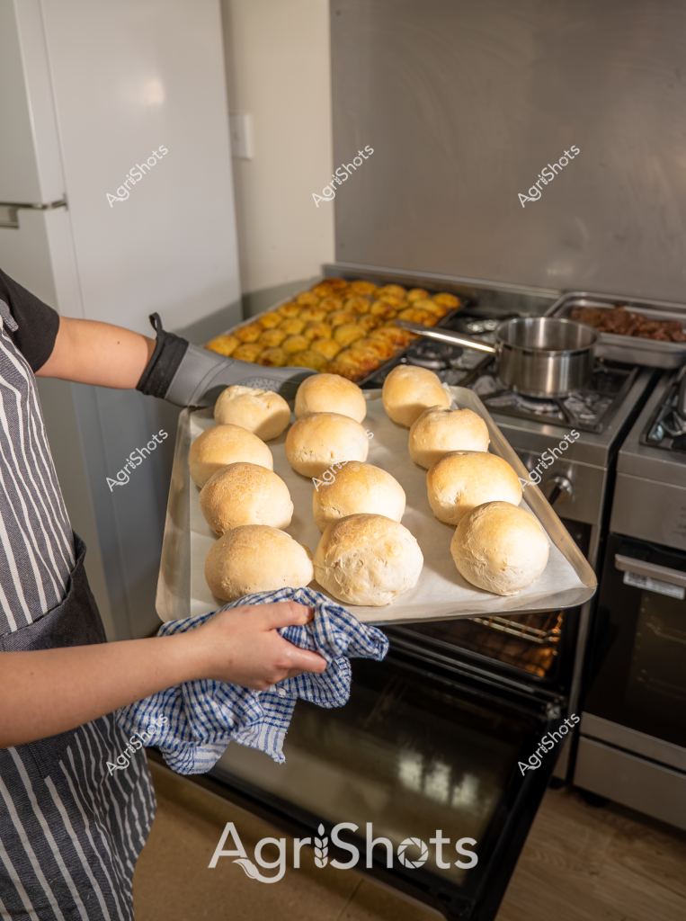 In this culinary scene, a woman in a blue and white striped apron carefully handles a baking tray filled with freshly prepared bread rolls. The golden-brown rolls are uniformly arranged, showcasing her skilled baking technique. In the background, a gas stove is visible with additional cooking trays, suggesting an active kitchen environment. A yellow tray of what appears to be roasted potatoes or another side dish sits near the stovetop, adding color and depth to the scene. The blue-and-white checkered kitchen towel in her hand emphasizes the traditional, homely atmosphere of bread-making. The warm, inviting colors and precise arrangement of the rolls highlight the precision and care involved in home baking, capturing a moment of culinary craftsmanship.
