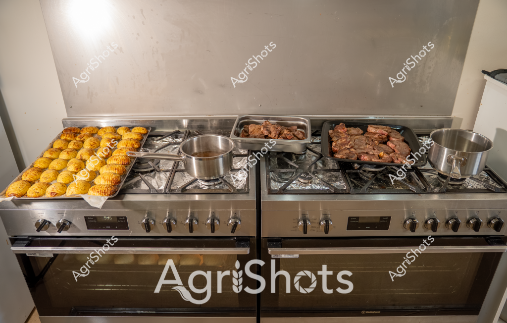 A professional kitchen scene captures a bustling culinary preparation, with two large stainless steel gas stoves filled with cooking activity. On the left side, rows of golden, freshly baked pastries - likely dinner rolls or small golden breads - are neatly arranged on a tray, their surfaces perfectly browned and glossy. Adjacent to the pastries, stainless steel pots sit on the stovetop, while on the right side of the stove, large cooking trays hold what appears to be pieces of roasted or grilled meat, their rich brown color indicating careful cooking and caramelization.

The stoves are professional-grade, with multiple burners and digital displays, suggesting a commercial or high-end home kitchen environment. Stainless steel dominates the color palette, reflecting a clean, professional cooking space. The cooking surfaces are covered with aluminum foil, a common practice for easy cleanup and even heat distribution in professional kitchens.

The image represents a moment of food preparation, with multiple dishes simultaneously in progress - baking, boiling, and roasting - capturing the dynamic energy of cooking. The careful arrangement and precise cooking techniques suggest a professional or highly skilled culinary setting, where attention to detail and technique are paramount.