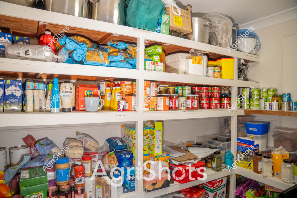 This image depicts a well-stocked pantry or kitchen storage area with multiple white shelves densely packed with a variety of food products, dry goods, and household supplies. Blue, red, and yellow packaging creates a colorful and organized display of groceries, including bags of pasta or grains, canned goods, jars of condiments, milk cartons, and various packaged food items. The shelves are completely full, suggesting a well-prepared household with a comprehensive food supply. Plastic storage containers, tea boxes, and other kitchen staples are strategically arranged to maximize the storage space, reflecting an efficient and practical approach to home food management.

The shelving system appears to be custom-built, with sturdy white wooden shelves extending from floor to ceiling, providing ample storage in what seems to be a dedicated pantry or storage area. The diverse range of products indicates a well-rounded food inventory, with everything from baking ingredients to canned vegetables, snacks, and beverages carefully organized and easily accessible.