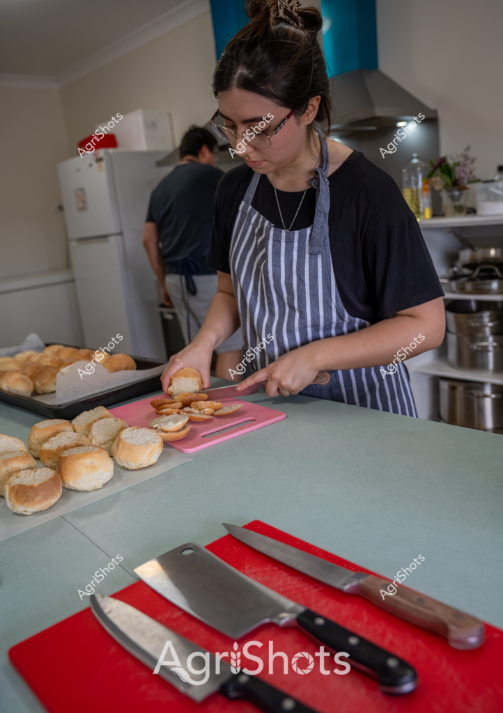 A woman wearing a blue and white striped apron and black t-shirt carefully prepares bread rolls in a commercial kitchen. She stands at a kitchen bench with a pink cutting board, methodically slicing freshly baked golden-brown bread rolls. Several additional rolls are arranged on baking trays nearby, suggesting a food preparation process. Sharp professional kitchen knives rest on a red cutting board in the foreground, indicating a professional food service or bakery setting. In the background, a white refrigerator and kitchen equipment can be seen, with another person partially visible, creating a sense of a busy, collaborative kitchen environment.

The scene captures the meticulous work of food preparation, with careful attention to detail evident in the woman's precise cutting technique and organized workspace. The neutral kitchen tones are punctuated by the vibrant red cutting board and the warm golden color of the freshly baked bread rolls, creating a visually appealing composition that speaks to professional food production.