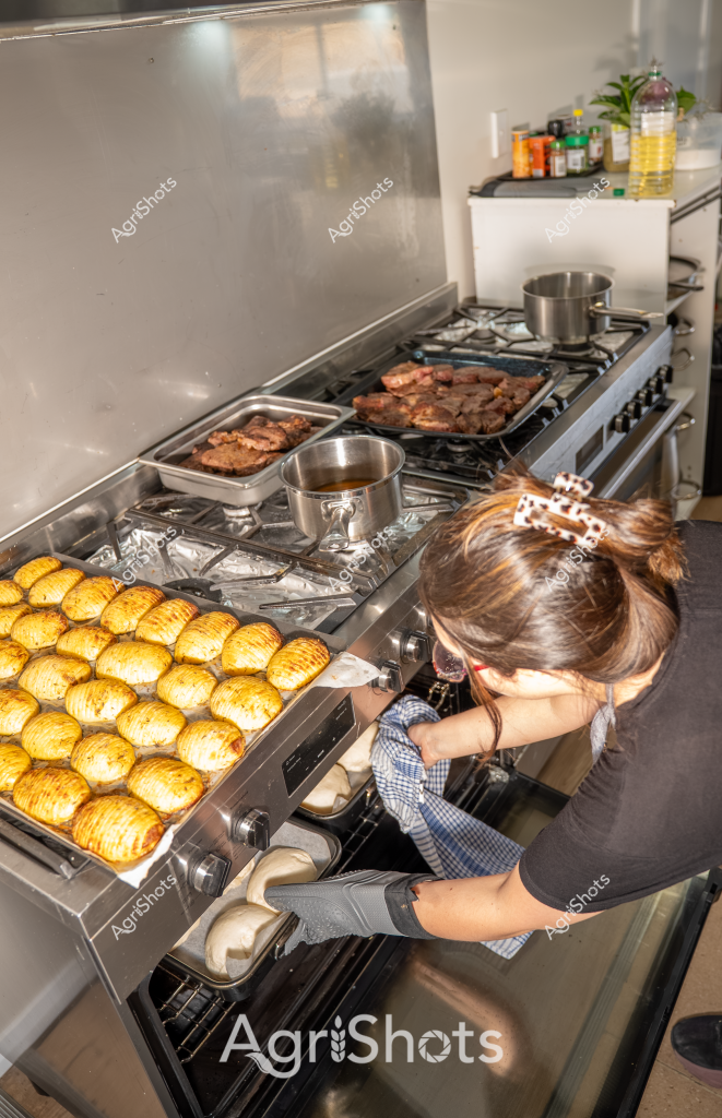 A professional kitchen scene captures a woman carefully preparing multiple dishes on a large stainless steel stove. Golden, precisely sliced potatoes are arranged meticulously across one section of the stovetop, their uniform rows suggesting careful culinary technique. Next to the potatoes, multiple trays of meat are being simultaneously cooked - appearing to be steaks or roasted cuts with a rich, caramelized brown color. The woman, wearing a dark top and protective kitchen gloves, is leaning over the stove, concentrating on her cooking process. Cooking vessels like stainless steel pots are strategically placed on the gas burners, indicating a professional and organized cooking environment. Bottles of cooking oils and ingredients are visible in the background, completing the mise en place of this busy kitchen preparation.

The image represents a moment of professional culinary craft, showcasing precision, multi-tasking, and attention to detail in food preparation. The golden potatoes and seared meats suggest a high-end restaurant or catering kitchen setting, with the woman demonstrating skilled cooking techniques.