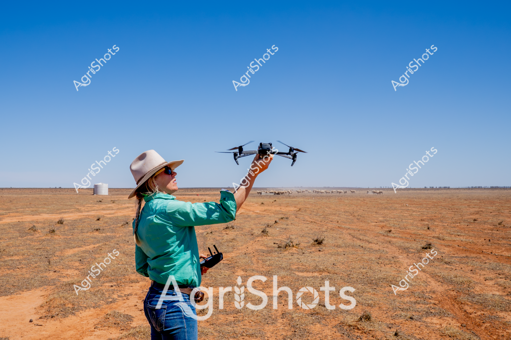 A woman in a teal shirt and wide-brimmed hat stands in a dry, expansive outback landscape, operating a drone for agricultural monitoring. The reddish-orange terrain stretches to a distant horizon, punctuated by sparse vegetation and a few water tanks in the background. She holds a drone controller while launching a quadcopter drone into the clear blue sky, demonstrating modern precision agriculture techniques. The arid environment suggests this is likely a remote cattle station or agricultural property in inland Australia, where technological solutions help farmers manage vast, challenging landscapes. Her professional stance and equipment highlight the innovative approaches used in contemporary Australian agricultural practices, bridging traditional farming with cutting-edge aerial surveying technology.