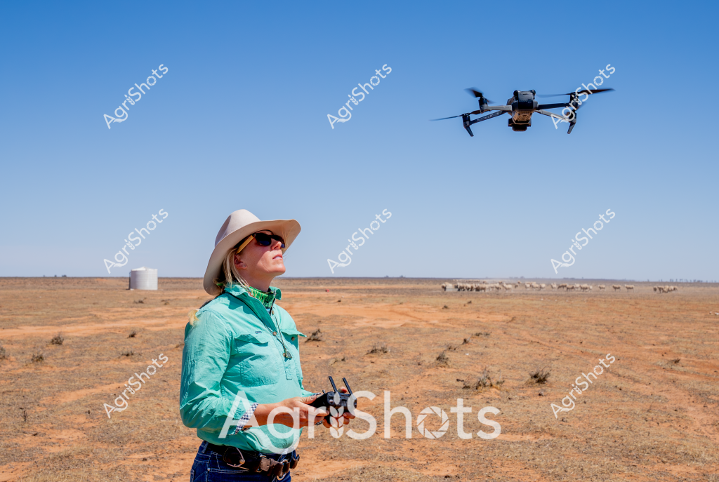 A woman in a turquoise shirt and wide-brimmed hat operates a drone in an arid, expansive Australian outback landscape. Standing confidently with a drone controller in her hands, she pilots an unmanned aerial vehicle (UAV) above a dry, reddish-brown paddock with scattered sparse vegetation. In the background, a distant water tank and a small herd of cattle can be seen, highlighting the remote agricultural setting. The clear blue sky and open terrain suggest this is a typical drone reconnaissance mission for modern farm management, likely used for monitoring livestock, assessing land conditions, or surveying crop and pasture health. Her protective clothing - including sunglasses and a wide-brimmed hat - demonstrates the harsh environmental conditions of rural Australian rangelands.