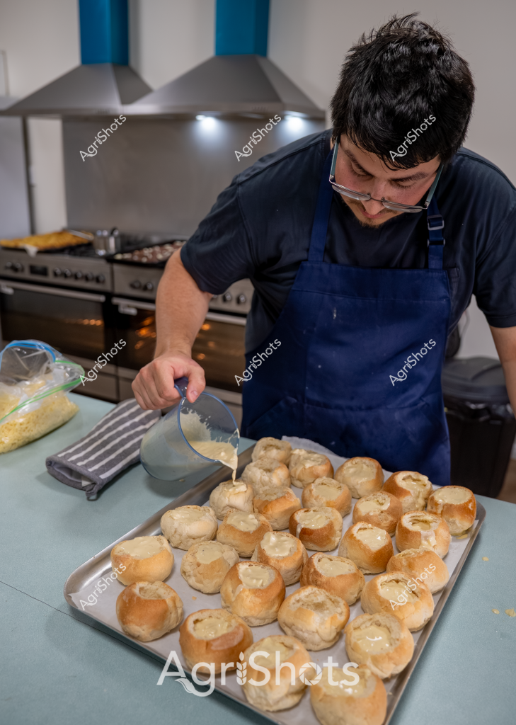 A young man wearing a blue apron and glasses carefully prepares a batch of golden-brown bread rolls in a professional kitchen setting. He is pouring a light-colored liquid, likely an egg wash or milk glaze, over a tray of freshly baked rolls with hollowed tops. The kitchen features stainless steel range hoods and a commercial stove in the background, indicating a professional baking environment. The bread rolls are uniformly shaped and arranged neatly on a large baking tray, showcasing precision and skill in bread preparation. The baker's focused expression and careful technique suggest he is creating artisan-style bread rolls, likely for a restaurant, bakery, or catering service.