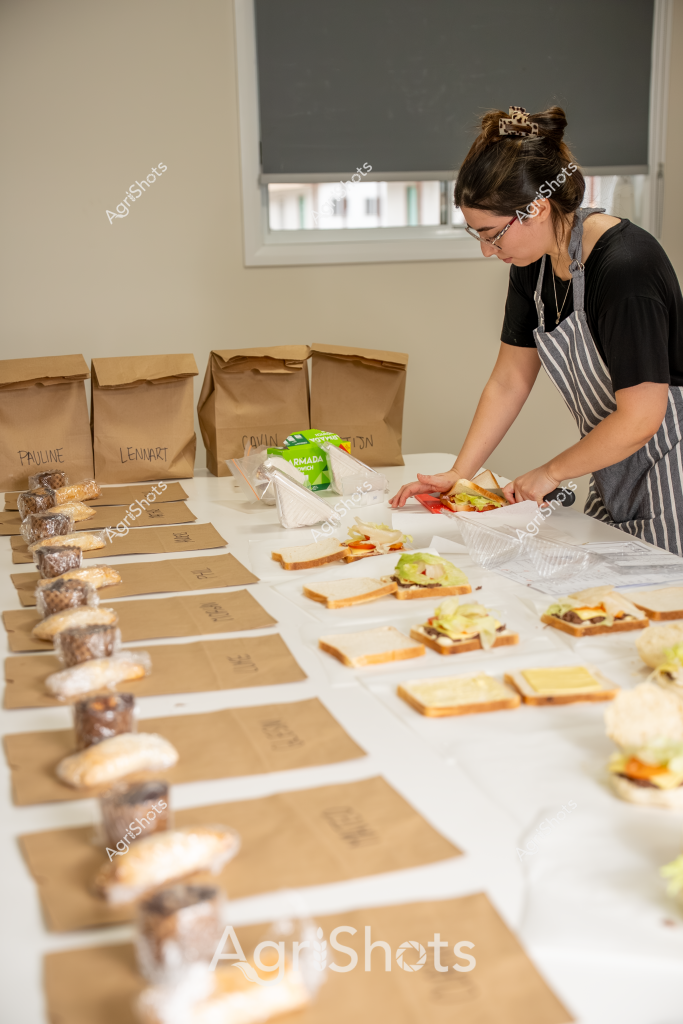 A woman in a striped apron and black t-shirt carefully prepares a series of individually labeled lunch bags and sandwiches in what appears to be a commercial food preparation space. Brown paper bags are meticulously arranged with names like "Pauline" and "Lennart" written on them, while an array of sandwich ingredients are spread out across the work surface. Several containers with baked or packaged items are positioned alongside the freshly prepared sandwiches, suggesting a systematic approach to meal preparation. The scene reflects precision and organization, with each component carefully placed and labeled, indicating this is likely a catering or meal delivery service operation. The neutral background with a pulled-down window shade creates a clean, focused environment for food preparation.