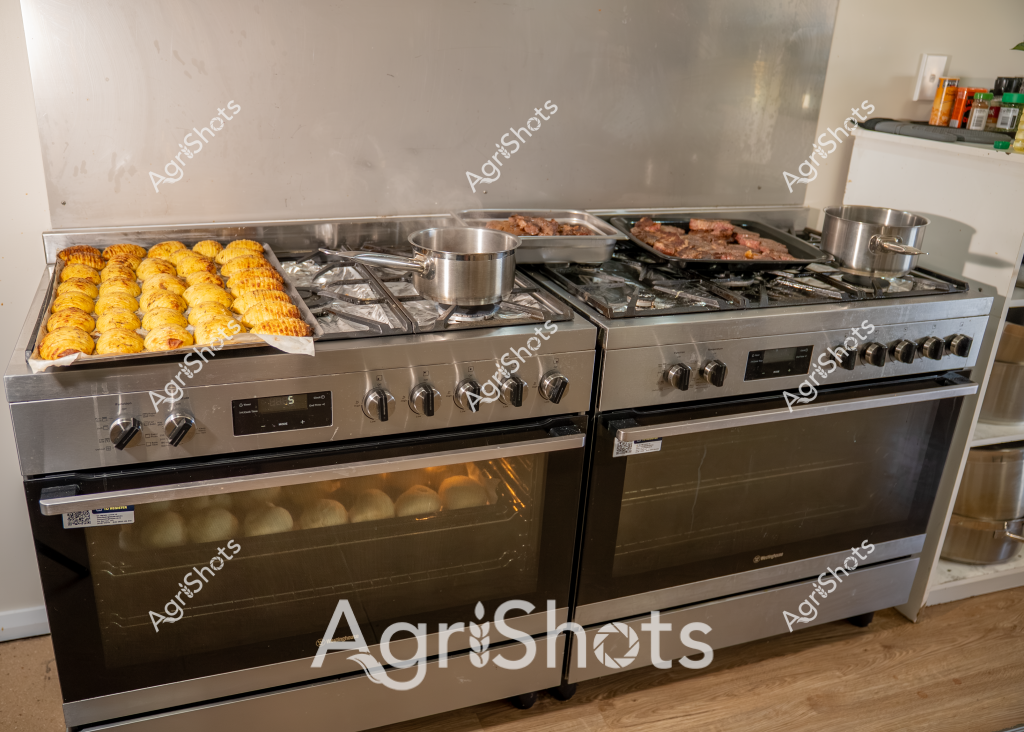 The image showcases a professional kitchen stove setup with multiple cooking elements in progress. Golden-colored pastries or bread rolls are neatly arranged on a baking tray on the left side of the stove, while a meat tray with what appears to be beef or steak is cooking on the right burner. Stainless steel pots are positioned on the stovetop, indicating an active cooking environment. The large stainless steel commercial-grade stove has multiple gas burners and digital control panels, suggesting a professional or high-end home kitchen setting. The oven below is partially visible, with additional round items visible through the glass door, hinting at ongoing baking preparations. The clean, organized layout and professional-grade equipment suggest a serious cooking or culinary preparation space.