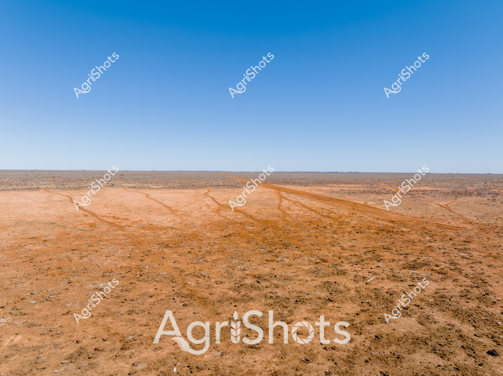 A vast, arid landscape stretches beneath an expansive blue sky, revealing a dry, reddish-orange terrain marked by faint vehicle tracks. The ground appears parched and barren, characteristic of drought-affected Australian outback regions, with rocky, sparse ground extending to a distant horizon. Multiple wheel tracks cut across the landscape, creating thin lines that snake through the dusty, sun-baked earth, suggesting recent vehicle movement across this remote and challenging terrain. The image captures the harsh environmental conditions typical of Australia's interior, where agricultural and pastoral activities must contend with extreme dry seasons and challenging landscape conditions. The stark contrast between the deep blue sky and the burnt-orange ground emphasizes the stark, unforgiving nature of this arid environment, highlighting the resilience required for survival and agricultural operations in such challenging regions.