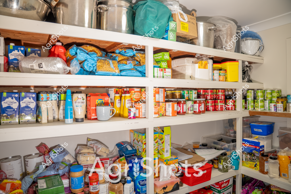 This image showcases a well-stocked pantry or kitchen storage area filled with a diverse array of food products and supplies. Multiple white shelves are densely packed with various packaged goods, including bags of dry ingredients, canned items, milk cartons, and assorted containers in blue, red, green, and yellow packaging. The shelves are meticulously organized, with items arranged by type and color, creating a visually appealing and practical storage system.

Blue bags of what appear to be dried goods are stacked alongside white milk cartons, yellow spice containers, and red-labeled items. The shelves extend from floor to ceiling, maximizing vertical storage space. Plastic containers, mixing bowls, and additional storage items are visible on the top shelf, indicating a comprehensive approach to kitchen organization.

The color palette is predominantly white, with pops of blue, red, green, and yellow from the various food packages. The shelving appears to be a combination of white-painted wooden shelves with some natural wood elements visible. The overall impression is one of preparedness, organization, and a well-stocked kitchen ready for meal preparation.