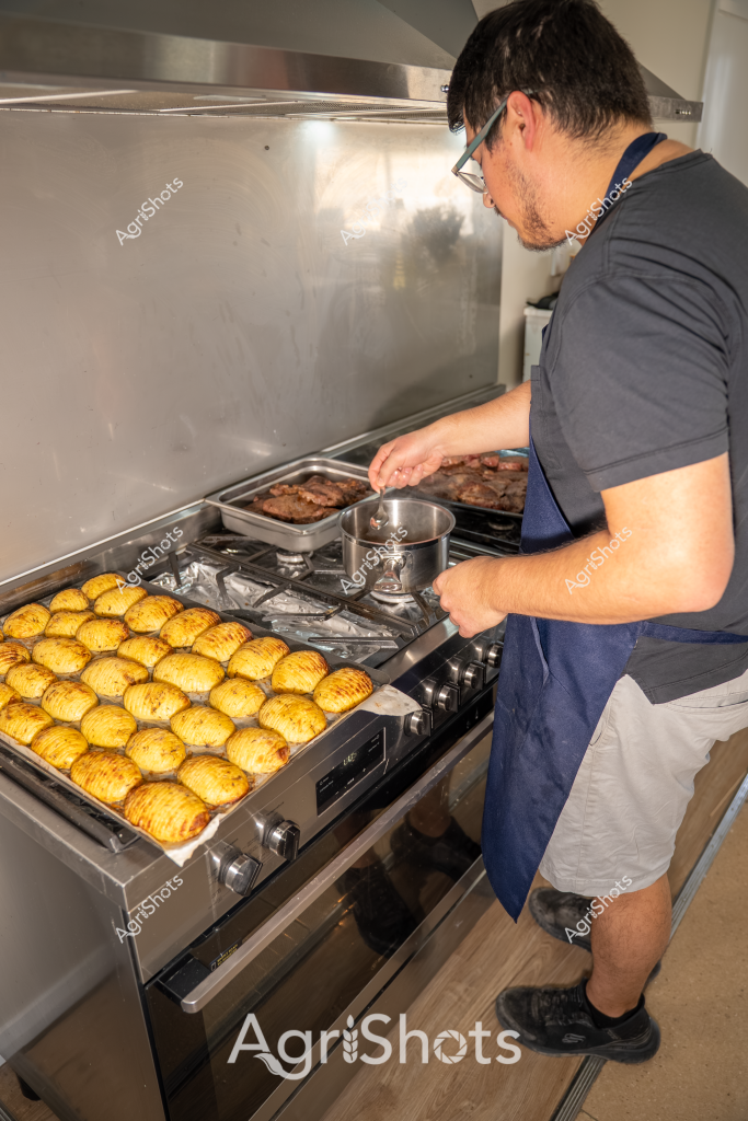 A man in a blue apron and gray t-shirt prepares a large batch of golden, perfectly roasted potatoes on a professional stainless steel stove. The potatoes are uniformly cut and browned, arranged meticulously across the stovetop in neat rows, suggesting careful culinary preparation. Simultaneously, he is cooking what appears to be meat in another pan, indicating a multi-tasking cooking process. The kitchen environment looks clean and professional, with a stainless steel backsplash and modern appliances. The golden color of the potatoes contrasts beautifully with the dark stove and the man's blue apron, creating a visually appealing scene of food preparation.

The image captures a moment of precise cooking technique, with the potatoes looking evenly roasted and glistening - likely seasoned and prepared with skill. The man's focused posture and careful positioning suggest he is an experienced cook or chef working methodically in a professional kitchen setting.