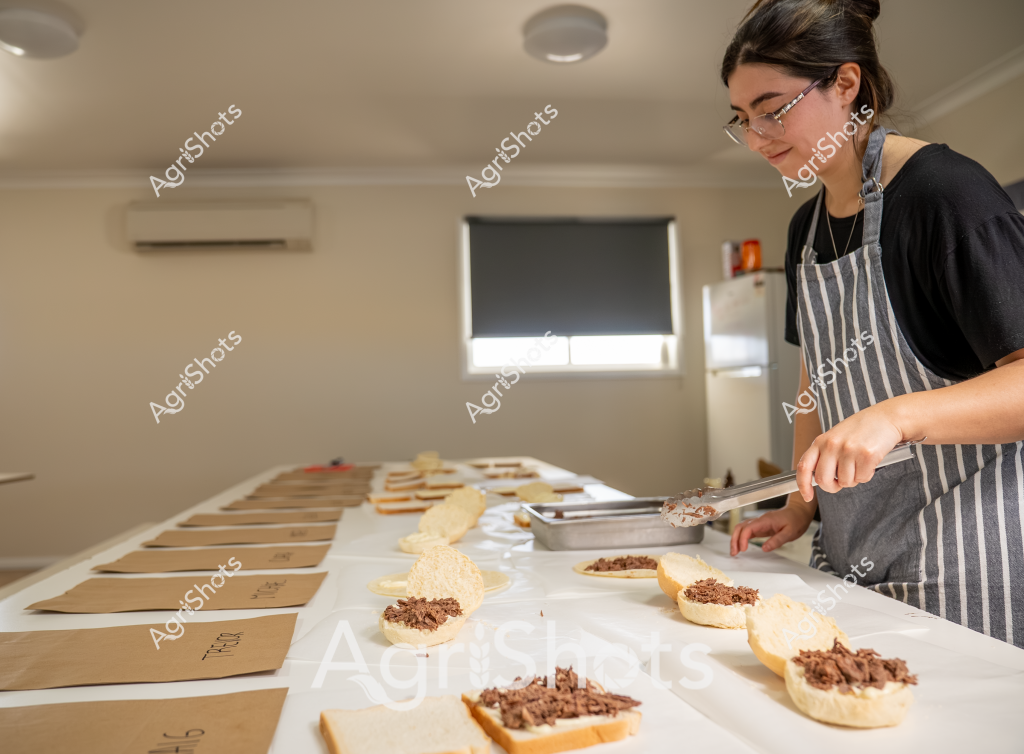 A woman wearing glasses and a striped apron carefully prepares a series of open-faced sandwiches in a commercial kitchen setting. She is methodically spooning what appears to be a ground meat filling onto white bread slices, with several prepared sandwiches already arranged on a white surface. Brown kraft paper bags are lined up on the left side of the workspace, suggesting these may be part of a catering or meal preparation process. The kitchen environment is clean and neutral-toned, with a window roller blind, air conditioning unit, and refrigerator visible in the background. Her focused expression and precise movements indicate professional food preparation techniques, with each sandwich being assembled with careful attention to detail.