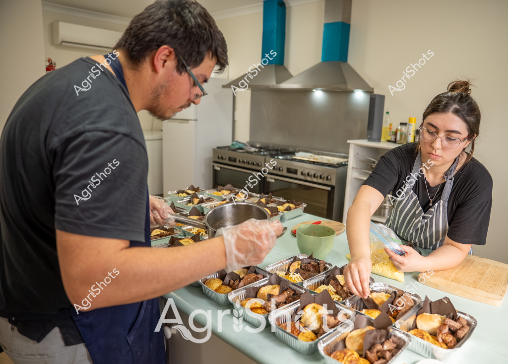 Two workers carefully prepare meal service trays in a commercial kitchen setting. A man wearing a dark shirt and protective gloves is working alongside a woman in a striped apron and glasses, arranging multiple aluminum takeaway containers filled with various cooked food items. The kitchen features a stainless steel stove and range hood in the background, with the workspace covered in neatly organized meal portions that appear to include meat, potatoes, and other prepared dishes. Both workers are focused and methodical, suggesting this is a professional catering or meal preparation operation, with precise attention to portioning and presentation.

The scene captures the behind-the-scenes work of food preparation, with multiple identical trays indicating this is likely a large-scale meal service for an institution, event, or delivery service. The careful placement of each item and the use of professional kitchen equipment highlights the systematic approach to creating consistent, portioned meals.