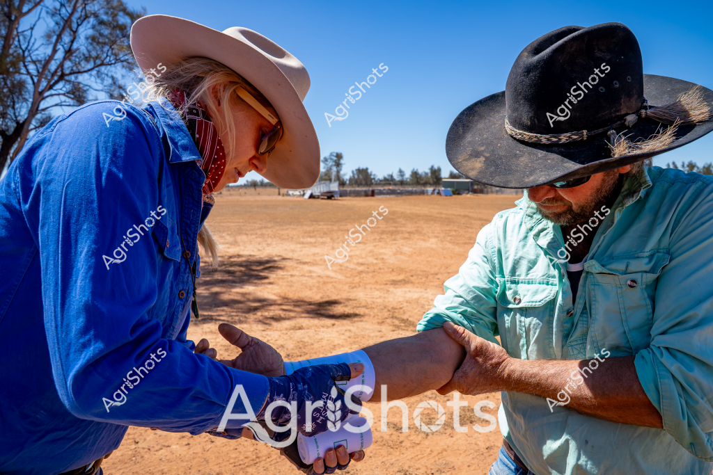 Two rural workers perform a medical check on an arm in an arid Australian landscape. A woman in a bright blue long-sleeved shirt and light-colored wide-brimmed hat carefully examines the arm of a man wearing a light blue shirt and a dark, well-worn wide-brimmed hat. The background reveals a dry, dusty paddock with sparse trees and farm infrastructure, indicating a remote station or rural property. The bright blue sky suggests a clear, hot day typical of the Australian outback. Their protective clothing, including long sleeves and wide-brimmed hats, demonstrates practical preparation for working in harsh environmental conditions. The careful, close interaction suggests a professional medical assessment or workplace injury check, highlighting the collaborative and supportive nature of rural work environments.