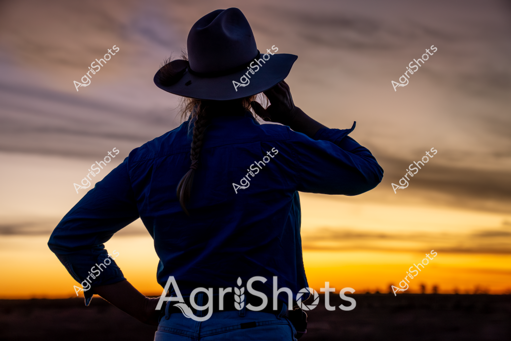 A woman in a wide-brimmed dark blue hat stands silhouetted against a dramatic sunset sky, facing away from the camera. Her blue jacket and braided hair are clearly defined against the golden-orange horizon, creating a striking visual of rural solitude and contemplation. The landscape stretches out behind her in soft, muted tones, with distant horizon lines suggesting an expansive agricultural setting. The dramatic lighting highlights the contours of her figure, casting a sense of quiet reflection and connection to the land. Her pose - with one hand on her hip - conveys a sense of strength and independence typical of rural Australian women.