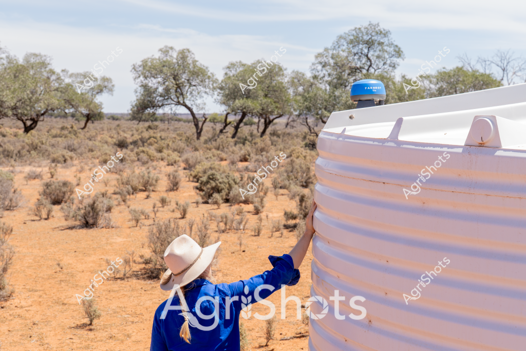 A woman wearing a blue shirt and a wide-brimmed sun hat stands beside a large white water tank in a dry, arid Australian landscape. Sparse trees and low scrubby bushes stretch across the dusty, reddish-brown terrain, typical of an inland pastoral region. The water tank appears to be a critical resource in this harsh environment, with a blue sensor or monitoring device mounted on its top. The scene captures the challenging conditions of rural Australian agriculture, where water storage is essential for survival in drought-prone areas. The woman's posture suggests she is checking or maintaining the water infrastructure, highlighting the ongoing management required in such remote and challenging landscapes.