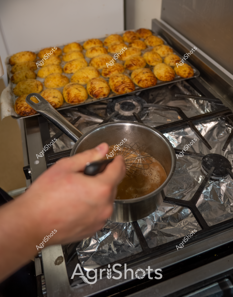 Here's a detailed description for this culinary scene:

Golden-brown potato gems radiate warmth across a baking tray, freshly roasted and clustered neatly on a gas stovetop. A hand wielding a stainless steel whisk stirs a small saucepan, suggesting a cooking technique involving preparing a complementary sauce or glaze. The potatoes appear crisp and uniformly golden, with a textured surface indicating they've been seasoned and roasted to achieve maximum crunchiness. The stove's aluminum foil-covered surface reflects the warm tones of the potatoes, creating a sense of home cooking and careful preparation. These appear to be classic potato gems or tater tots - a beloved side dish that promises a satisfying crispy exterior and soft interior, likely being prepared in a home kitchen with attention to culinary detail.

The image captures the precise moment of finishing a batch of perfectly roasted potato side dishes, with the cook actively engaged in the final stages of preparation.