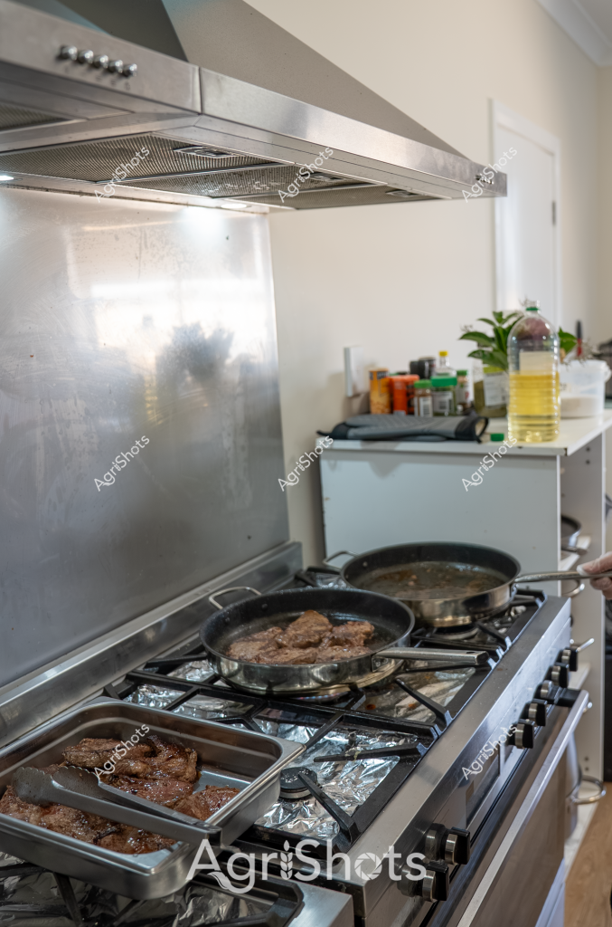 A kitchen scene captures meat cooking on a stainless steel gas stove, with multiple skillets and cooking trays searing what appears to be beef cuts. A sleek stainless steel range hood hangs overhead, with a metallic backsplash reflecting soft light. Cooking oil in a clear bottle and various spice containers are visible on a nearby countertop, suggesting a home cooking preparation. The meat looks well-seasoned, with a rich brown sear developing in the cast iron and stainless steel pans, indicating careful culinary technique. Kitchen elements like the clean white walls, modern appliances, and organized cooking space suggest a contemporary home cooking environment.