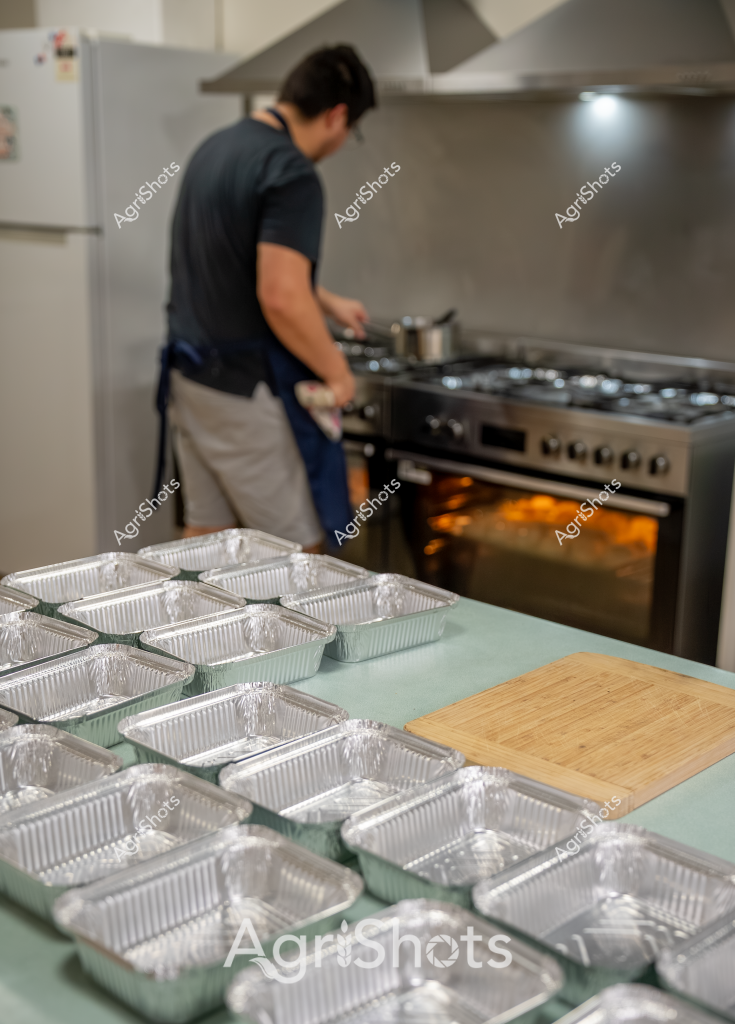 A professional kitchen scene captures a man in a dark shirt preparing multiple meal portions, with numerous aluminum takeaway containers lined up on a mint green countertop. An industrial-grade stove with an illuminated oven is visible in the background, suggesting a commercial food preparation environment. The aluminum containers are uniformly arranged, indicating a systematic approach to meal packaging or catering service. The kitchen appears clean and organized, with a wooden cutting board positioned nearby, hinting at a professional food service operation. The soft focus and careful arrangement suggest precision and attention to detail in food production, potentially for a meal delivery, catering, or restaurant takeaway service.