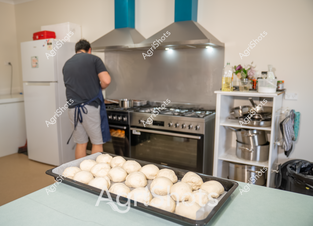 Here's a detailed description focusing on the culinary and kitchen scene:

A man wearing a dark grey t-shirt and grey shorts stands with his back turned in a commercial-style kitchen, preparing what appears to be bread or pastry. In the foreground, a large baking tray is filled with numerous round, raw dough balls - smooth, pale, and evenly shaped, suggesting preparation for bread or rolls. The kitchen features a stainless steel stove with a large exhaust hood, a white refrigerator, and a shelving unit with cooking equipment. The workspace is clean and organized, with cooking oils and ingredients visible in the background. The soft lighting and neutral color palette create a calm, professional atmosphere typical of a commercial or home bakery setting. The precise arrangement of the dough balls and the methodical positioning of the baker suggest careful preparation and attention to detail in the bread-making process.