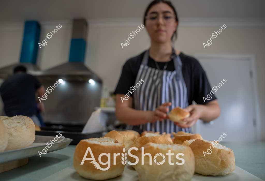 A woman baker wearing a black shirt and blue-and-white striped apron prepares freshly baked bread rolls in a commercial kitchen. Golden-brown bread rolls are arranged on a white surface in the foreground, their smooth crusts and rounded shapes suggesting recent baking. In the background, a commercial oven and kitchen equipment are partially visible, creating a professional food preparation environment. The baker stands focused, holding a bread roll and working with careful concentration. The scene captures the artisanal process of bread making, highlighting the precision and skill involved in creating quality baked goods.

The soft lighting and clean kitchen setting emphasize the professional nature of the food production, showcasing the care and attention that goes into traditional bread baking. The warm tones of the bread rolls contrast nicely with the cool kitchen environment, creating a visually appealing composition that speaks to culinary craftsmanship.