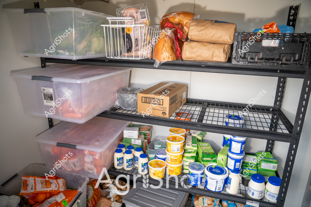 Based on the image provided, this appears to be a storage shelving unit in a food storage or pantry area, filled with various food products and storage containers. The shelves are industrial-style metal wire shelving, loaded with clear plastic storage bins, cardboard boxes, bagged goods, and assorted food containers in blue, white, and green packaging. The storage system looks organized, with items neatly arranged across multiple levels - from bread and packaged goods on the top shelf to dairy and other containers on the lower shelves. Several large clear plastic containers with lids are visible, suggesting this is a systematic food storage setup, likely in a commercial or large-scale kitchen or food preparation environment. The arrangement indicates careful inventory management and efficient use of vertical storage space.