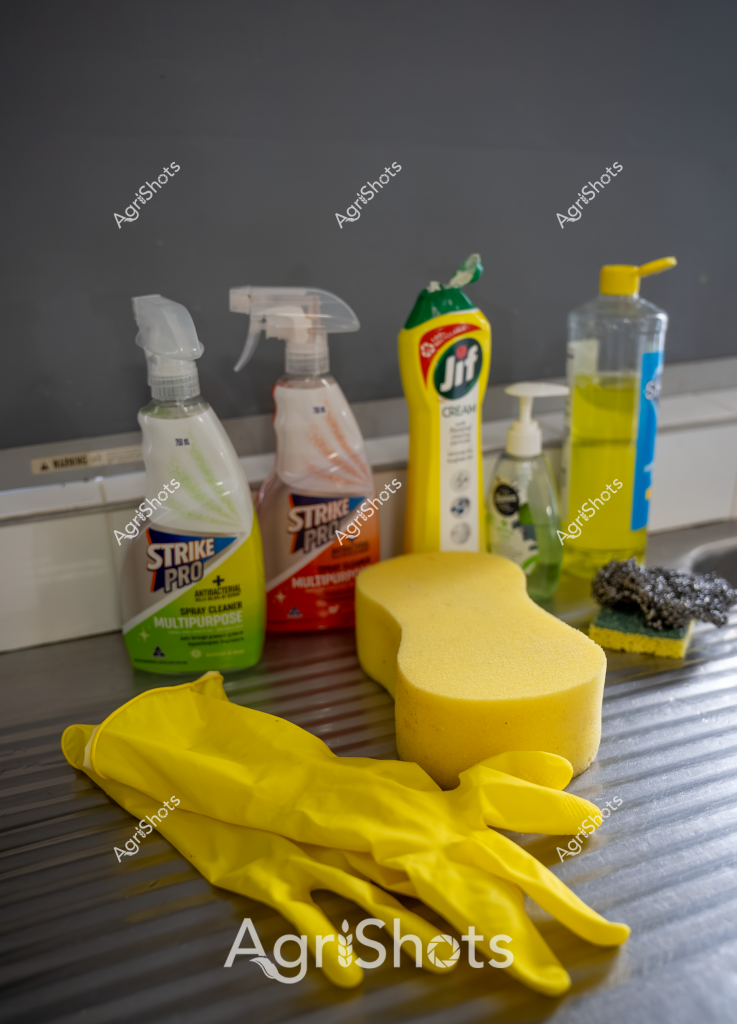 This image depicts a cleaning and home maintenance setup, featuring various cleaning products and supplies arranged on a surface. A pair of bright yellow rubber gloves dominates the foreground, alongside a large yellow cleaning sponge. Multiple spray bottles from brands like Strike Pro and Jif are positioned behind the gloves, indicating a range of cleaning solutions for different purposes. A steel wool scrubber and additional cleaning bottles complete the arrangement, suggesting a comprehensive cleaning kit ready for household or industrial cleaning tasks. The image has a professional, organized feel, with the bright yellow accessories creating a sense of preparedness and efficiency in tackling cleaning challenges.