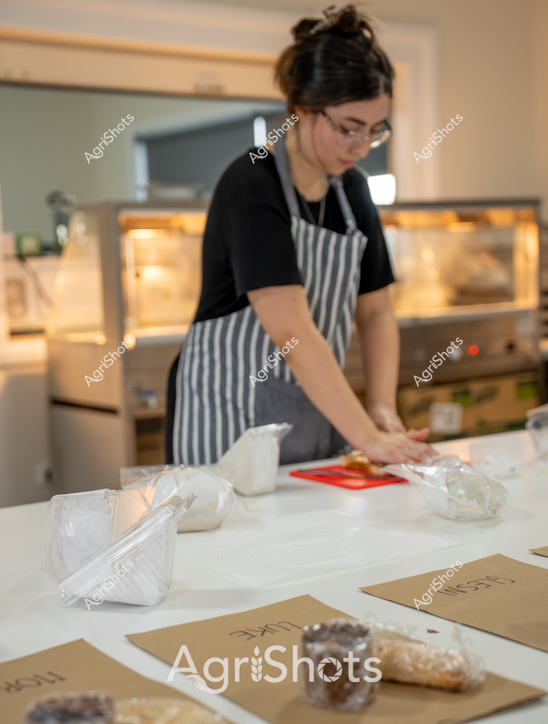 A woman wearing glasses and a black shirt with a gray and white striped apron works diligently in what appears to be a food preparation area. She is focused on preparing ingredients on a clean white countertop, surrounded by various packaging materials and kraft paper work surfaces. Clear plastic containers and bags are neatly arranged around her workspace, suggesting a professional food production or packaging environment. The background reveals warm-toned bakery or kitchen equipment, creating a soft, inviting atmosphere. Her hair is pulled back in a practical updo, and her careful movements indicate a methodical approach to her work, reflecting the precision required in food preparation and packaging.