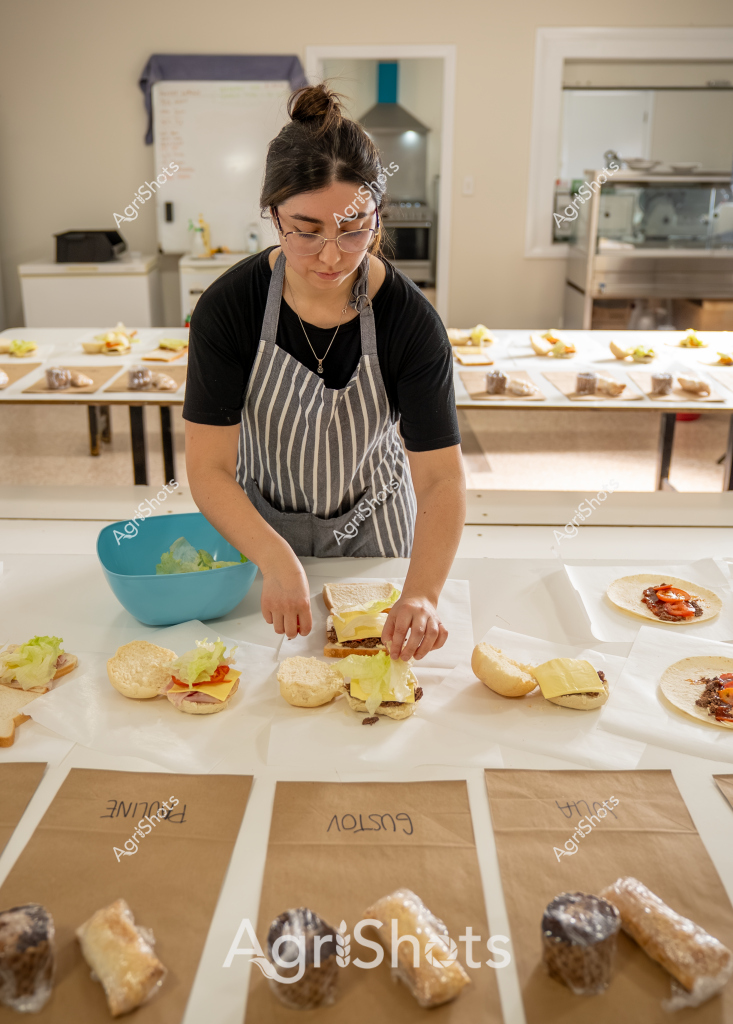 A woman in a gray striped apron and black t-shirt prepares multiple sandwiches in a professional kitchen setting. She carefully assembles burger components on white paper, with various ingredients and prepared rolls spread across her workstation. A blue mixing bowl containing lettuce sits nearby, and brown paper bags with handwritten labels are positioned in the foreground, suggesting a systematic food preparation process. The kitchen appears clean and organized, with stainless steel equipment visible in the background. Her focused demeanor and precise arrangement of ingredients indicate she is likely preparing a batch of sandwiches for catering, a restaurant service, or a commercial food preparation environment.