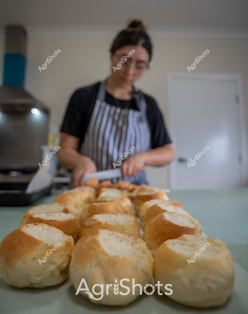 A woman baker stands in a kitchen, wearing a black t-shirt and blue-and-white striped apron, preparing freshly baked bread rolls. The golden-brown bread rolls are arranged neatly on a light-colored countertop, showcasing their perfectly crisp crusts and uniform shape. Her hands are positioned near the bread, suggesting she is in the process of arranging or preparing the baked goods. The background reveals a home kitchen setting with soft, neutral tones and a glimpse of kitchen equipment. The image captures a moment of culinary craftsmanship, highlighting the artisanal process of bread-making with its warm, inviting aesthetic.