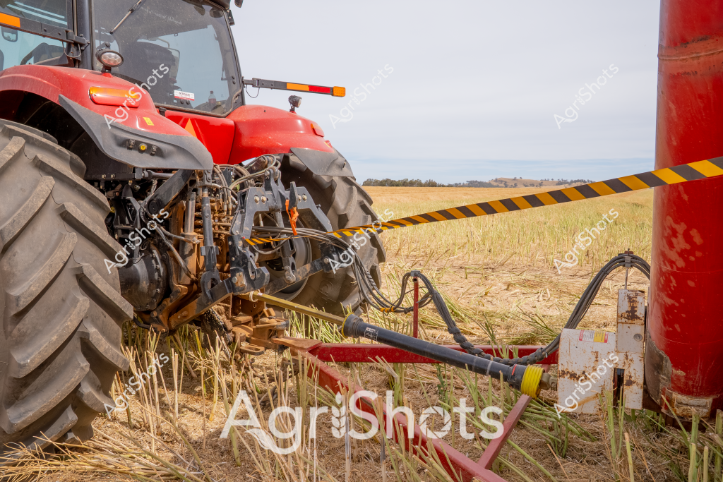 A robust red agricultural tractor stands in a harvested field, its large aggressive tread tires gripping the stubble-covered ground. Complex hydraulic linkages and mechanical connections are clearly visible between the tractor's rear attachment points, revealing sophisticated agricultural engineering. The tractor appears to be positioned for field work, with black hydraulic hoses connecting various mechanical components and a warning tape barrier visible in the background. Dry grass stubble surrounds the machinery, suggesting this is a post-harvest agricultural setting with expansive open paddock landscapes visible in the distance. The gleaming red paint of the tractor contrasts sharply with the muted earth tones of the surrounding agricultural landscape, highlighting modern farming equipment's sophisticated design and functionality.