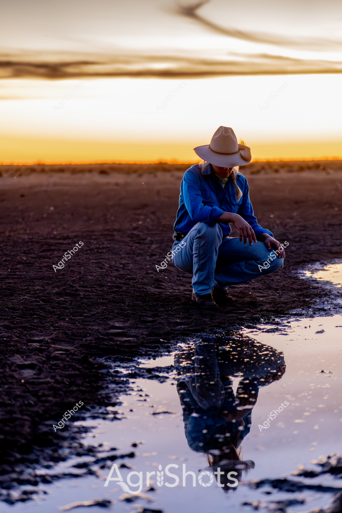 A solitary figure crouches at sunset in a stark, reflective landscape, embodying a moment of quiet contemplation. Wearing a classic wide-brimmed cowboy hat and a vivid blue work shirt, the person sits low to the ground, their reflection perfectly mirrored in a shallow pool of water. The background reveals a wide, flat horizon bathed in golden-orange light, characteristic of the Australian outback at dusk. Dark, textured ground surrounds the water's edge, creating a dramatic contrast with the luminous sky. The scene captures a profound sense of solitude and connection to the landscape, suggesting the introspective nature of rural life and the intimate relationship between a person and their environment.

The composition is striking in its simplicity - a single human form against an expansive, minimalist backdrop. The reflection creates a symmetrical visual poetry, with the figure seeming to exist in two planes simultaneously - both physically present and dreamlike in their watery double. This image could easily represent the resilience, isolation, and quiet strength associated with Australian rural and agricultural experience.