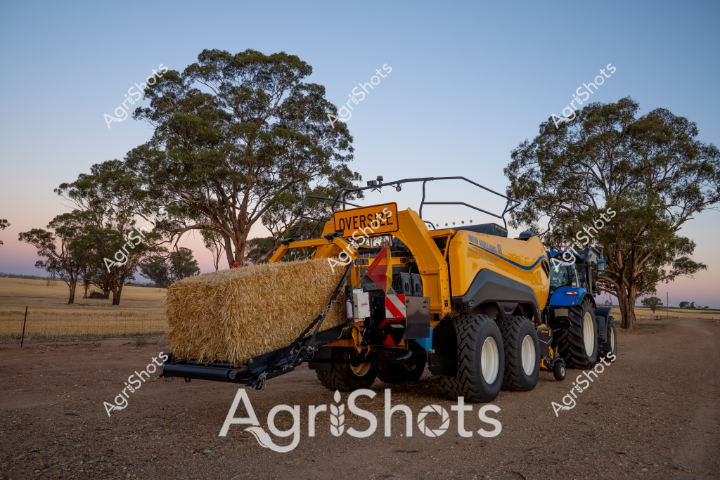 A gleaming yellow New Holland large square baler captures the essence of modern agricultural machinery, positioned against a serene rural landscape of scattered eucalyptus trees at dawn or dusk. The powerful piece of farm equipment is handling a large golden straw bale, demonstrating the precision of contemporary hay harvesting technologies. Crisp white wheels and intricate mechanical components contrast with the bright yellow body of the baler, highlighting the machine's robust design for efficient agricultural operations. The surrounding landscape features a classic Australian farm setting - dry paddocks with sparse trees and a soft, pastel sky creating a tranquil backdrop to the powerful farming equipment. The scene epitomizes the intersection of advanced agricultural technology and the timeless Australian rural environment, showcasing the sophisticated machinery that enables modern farming practices.

The baler appears to be parked on a dirt area, likely a farm yard or access road, with native trees framing the background. Its large square bale-handling capability represents the high-efficiency standards of contemporary agricultural equipment, designed to maximize productivity in challenging rural conditions.
