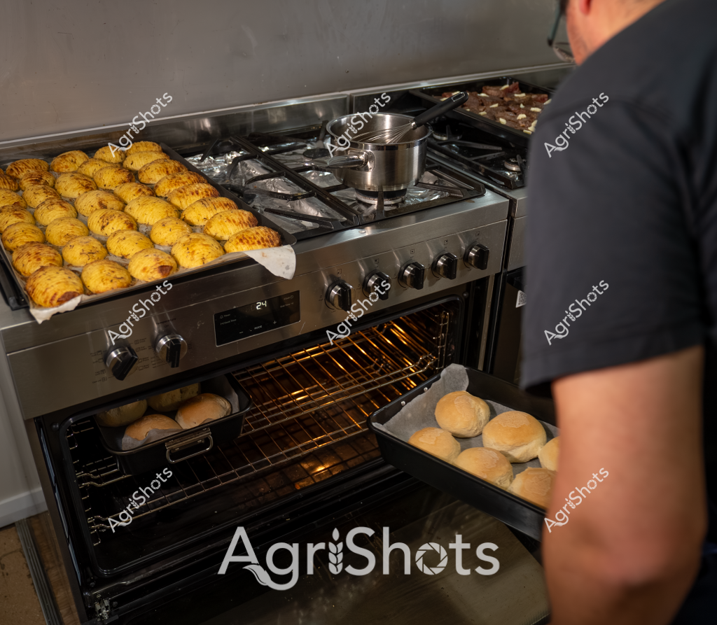 A professional kitchen scene showcases golden-brown pastries being prepared, with a stainless steel stove loaded with baked goods. Rows of perfectly uniform golden potatoes or rolls occupy the stovetop and oven racks, suggesting a bakery or commercial cooking environment. A stainless steel pot sits on the gas burner, while a person in a dark shirt stands partially visible, indicating ongoing food preparation. The oven's warm interior and neatly arranged baked items suggest a systematic, precise approach to cooking, with multiple trays of golden-colored baked goods at different stages of preparation. The gleaming appliance and organized layout highlight the methodical nature of professional food production, with each item carefully positioned for optimal cooking.
