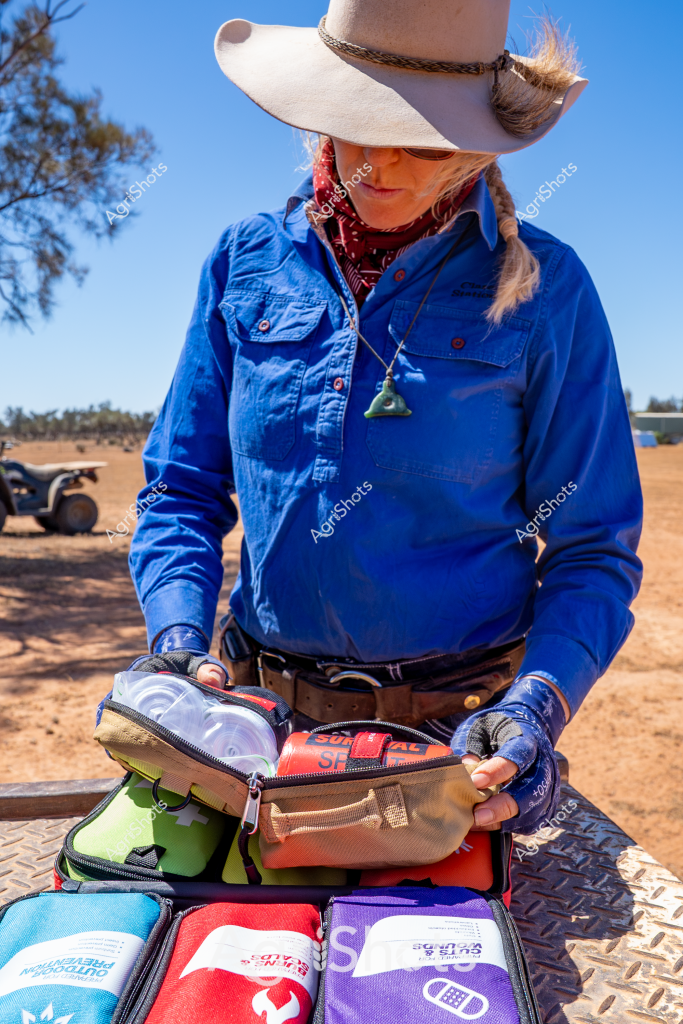 A woman wearing a wide-brimmed hat and blue work shirt carefully organizes a first aid or emergency medical kit on what appears to be a rural equipment surface. Her red bandana and agricultural-style work attire suggest she is a professional working in a remote or farm environment, likely preparing medical supplies for potential workplace or livestock management needs. The kit contains multiple colored pouches in red, white, purple, and blue, indicating a comprehensive emergency response package designed for rural or agricultural settings. The background reveals a dusty, open landscape with hints of farm equipment and trees, typical of an Australian agricultural working environment. Her gloved hands and focused posture demonstrate professional preparedness and attention to critical safety protocols in potentially challenging outdoor work conditions.

The scene captures the essential medical readiness required in remote agricultural workplaces, where quick access to emergency supplies can be crucial for worker and animal safety. The carefully organized medical kit reflects the methodical and practical approach characteristic of Australian rural professionals.