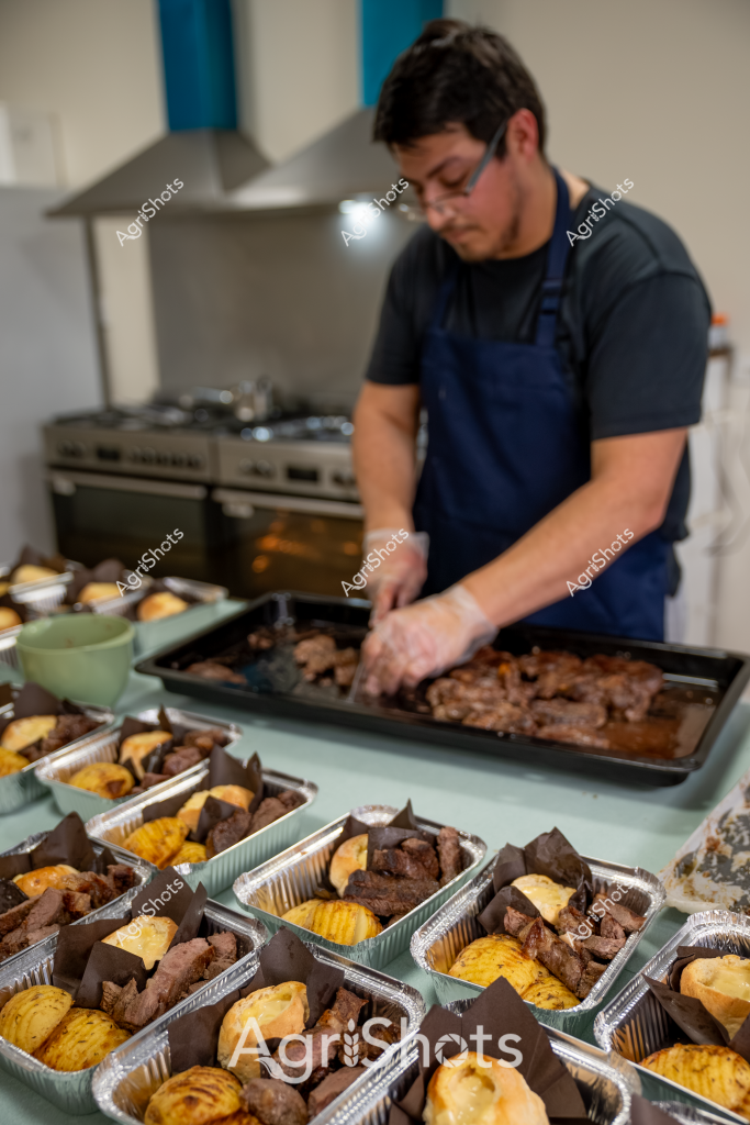 A professional kitchen scene shows a man in a dark t-shirt and navy blue apron preparing multiple pre-packaged meal portions. Aluminum trays are filled with what appears to be braised meat alongside golden-yellow baked items, likely a combination of roasted potatoes or cornbread. The man is carefully arranging or preparing these meal containers on a stainless steel kitchen counter, with a commercial stove visible in the background. Each tray contains a carefully portioned meal with dark meat cuts and complementary golden side dishes, suggesting a professional catering or meal preparation operation. The image captures the precision and methodical approach of commercial food service, with multiple identical meal portions meticulously arranged and ready for distribution.