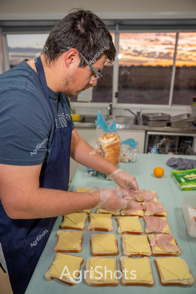 A man in a navy blue shirt and disposable gloves is preparing sandwiches in a commercial kitchen, carefully assembling multiple slices of bread with cheese and ham. The work surface is covered with neatly arranged toast slices, each layered with a slice of yellow cheese and a piece of ham. The kitchen has a sunset-lit window in the background, creating a warm glow and suggesting an early evening food preparation setting. He is wearing glasses and appears focused on his task, systematically constructing multiple sandwich bases with precision. Clear plastic food preparation gloves protect the food as he works, indicating professional food handling practices. A yellow citrus fruit and a stack of bread are visible nearby, suggesting additional ingredients for the meal preparation process.