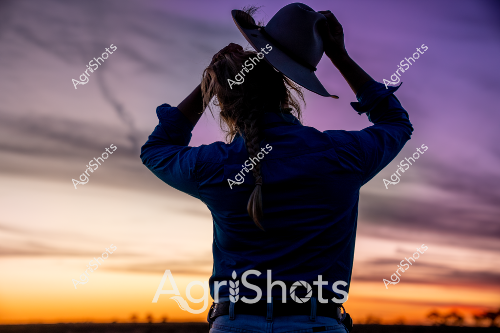 A woman wearing a blue jacket stands with her back to the camera, silhouetted against a dramatic sunset sky. She is holding a wide-brimmed hat above her head, revealing long hair pulled back in a ponytail. The background glows with warm orange and purple hues, creating a stunning gradient from the bright horizon to the deepening twilight. Soft clouds stretch across the sky, adding texture to the tranquil evening landscape. The scene captures a moment of quiet reflection, with the woman appearing to pause and appreciate the breathtaking rural sunset before her.