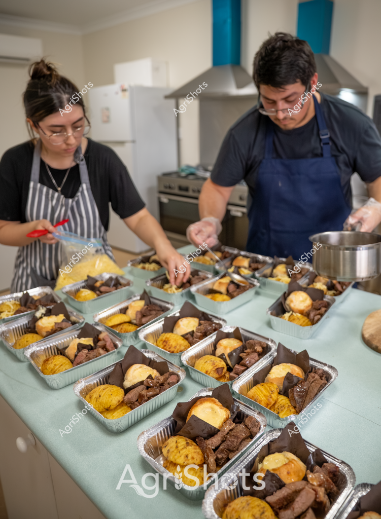 Two food service workers carefully prepare multiple meal portions in an industrial kitchen, arranging identical aluminum takeaway containers with precise culinary skill. A woman in a striped apron and a man in a navy blue apron are methodically plating identical meals, which appear to include seasoned yellow corn, dark meat portions, and golden-brown baked items. The kitchen environment is clean and professional, with stainless steel equipment and a white refrigerator visible in the background. Each aluminum container is uniformly filled with a consistent arrangement of food components, suggesting a professional catering or meal preparation service operation. The workers demonstrate careful attention to detail, ensuring each meal portion is identically composed and presented with precision.

The image captures the systematic and organized approach of commercial food preparation, with the two workers focused intently on their tasks of portioning and plating. Their coordinated movements and matching aprons indicate a well-trained team working efficiently to produce multiple identical meal servings.