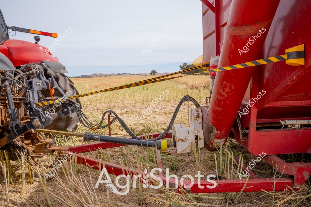 A red agricultural harvester and tractor are connected by hydraulic lines in a dry stubble field, showcasing modern farming equipment during harvest season. Distinctive black and yellow caution tape stretches between the machinery's connection points, highlighting safety protocols in agricultural operations. The golden-brown wheat or grain stubble covers the ground, indicating a recently harvested paddock with a sparse landscape of distant trees and open horizon. Mechanical components of the tractor reveal complex hydraulic systems and robust agricultural engineering, with the bright red paint of the machinery standing out against the muted landscape. Flexible hydraulic hoses curve between the machines, demonstrating the intricate connections required for efficient modern farming techniques. The scene captures the intersection of technological precision and agricultural landscape, reflecting the sophisticated approach to crop harvesting in contemporary farming.